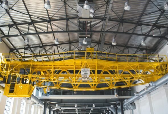 A yellow gantry crane inside an industrial factory with metal roof and lights.